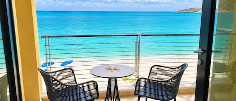 A balcony with two chairs and a table, overlooking a beach with clear turquoise water and a distant island.