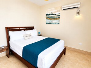 A neatly made bed with a blue blanket, a wall-mounted air conditioner, and a painting above the bed.