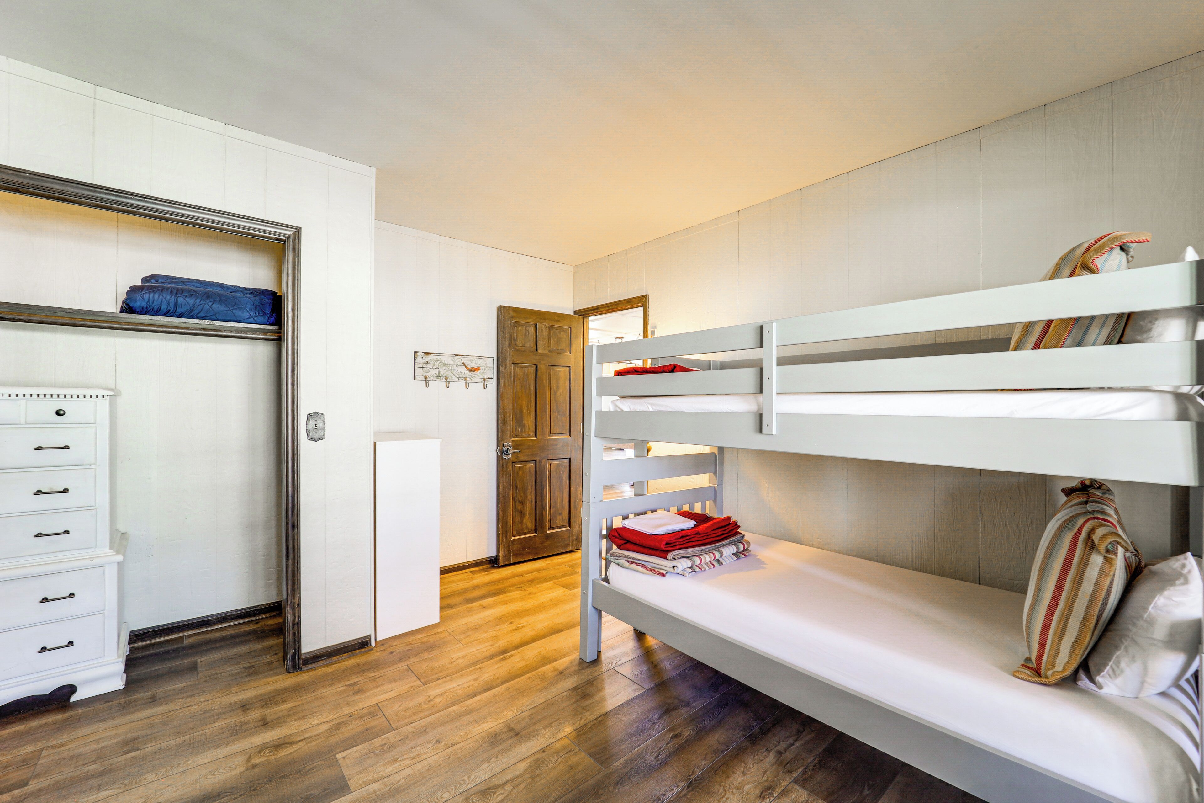 A bunk bed room with a wooden floor, a white dresser, and a door leading to another room.