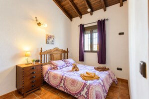 A bedroom with a wooden bed, a nightstand, a lamp, a window with curtains, and a wall-mounted light fixture. - Cubo's Casa La Viña / private pool / air conditioning / free wifi (Alhaurín el Grande)