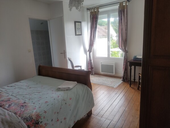 Un dormitorio con cama, una silla, una mesita, un ventanal con cortinas y una puerta que da a otra habitación.