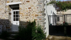 Ein Gebäude aus Stein mit einer weißen Tür und einem Fenster, umgeben von Grünpflanzen und einem schwarzen Tor.