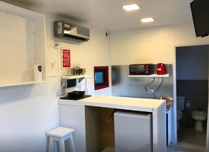 Una cocina compacta con microondas, tostadora y un refrigerador pequeño. Hay un taburete blanco y un cuadro con marco rojo en la pared.