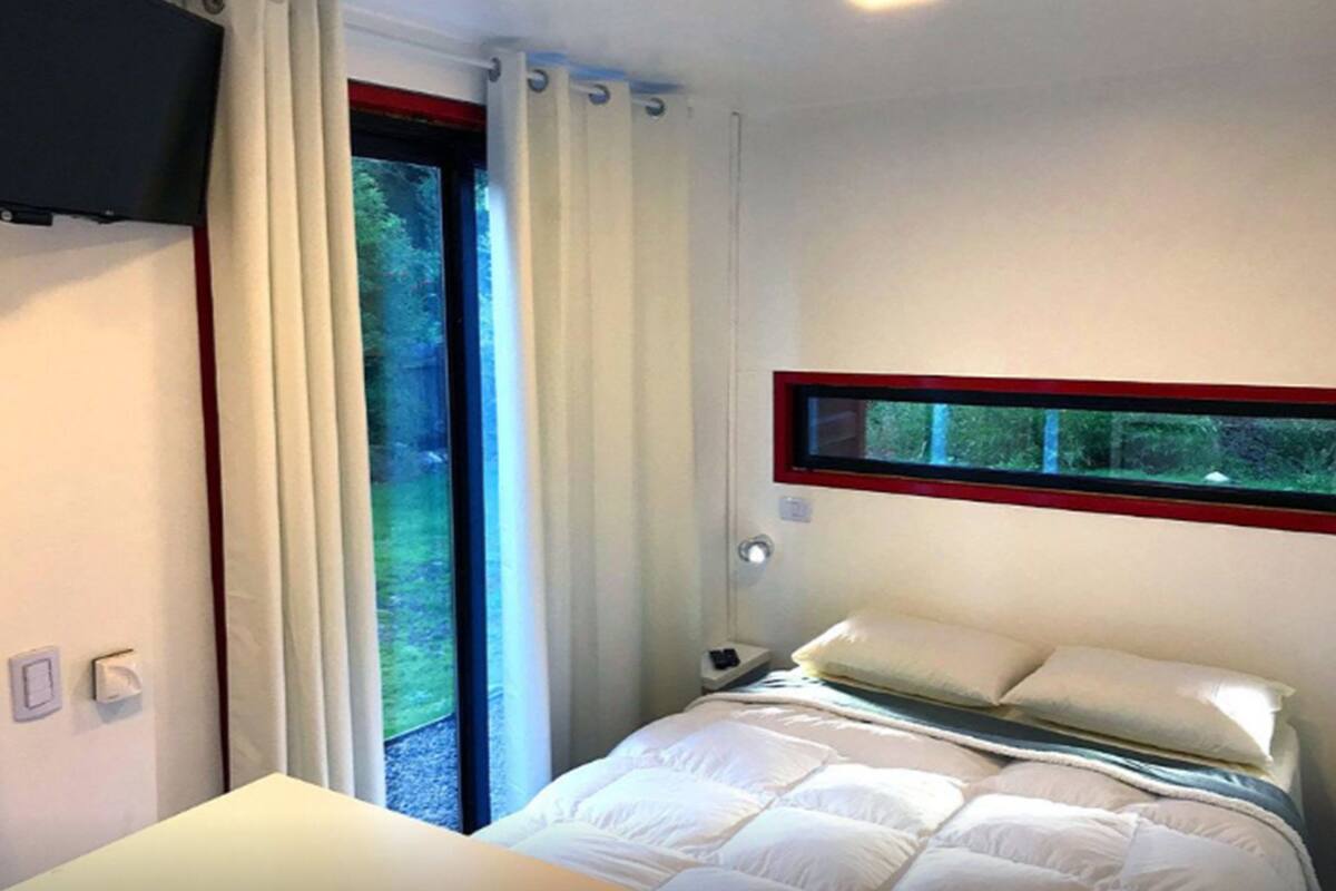 A bedroom with a bed, a window with curtains, a bedside table, and a wall-mounted TV.