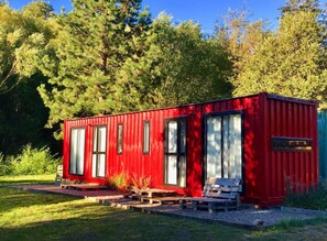 Una casa contenedor roja con ventanas grandes, un banco de madera y un sendero de grava.