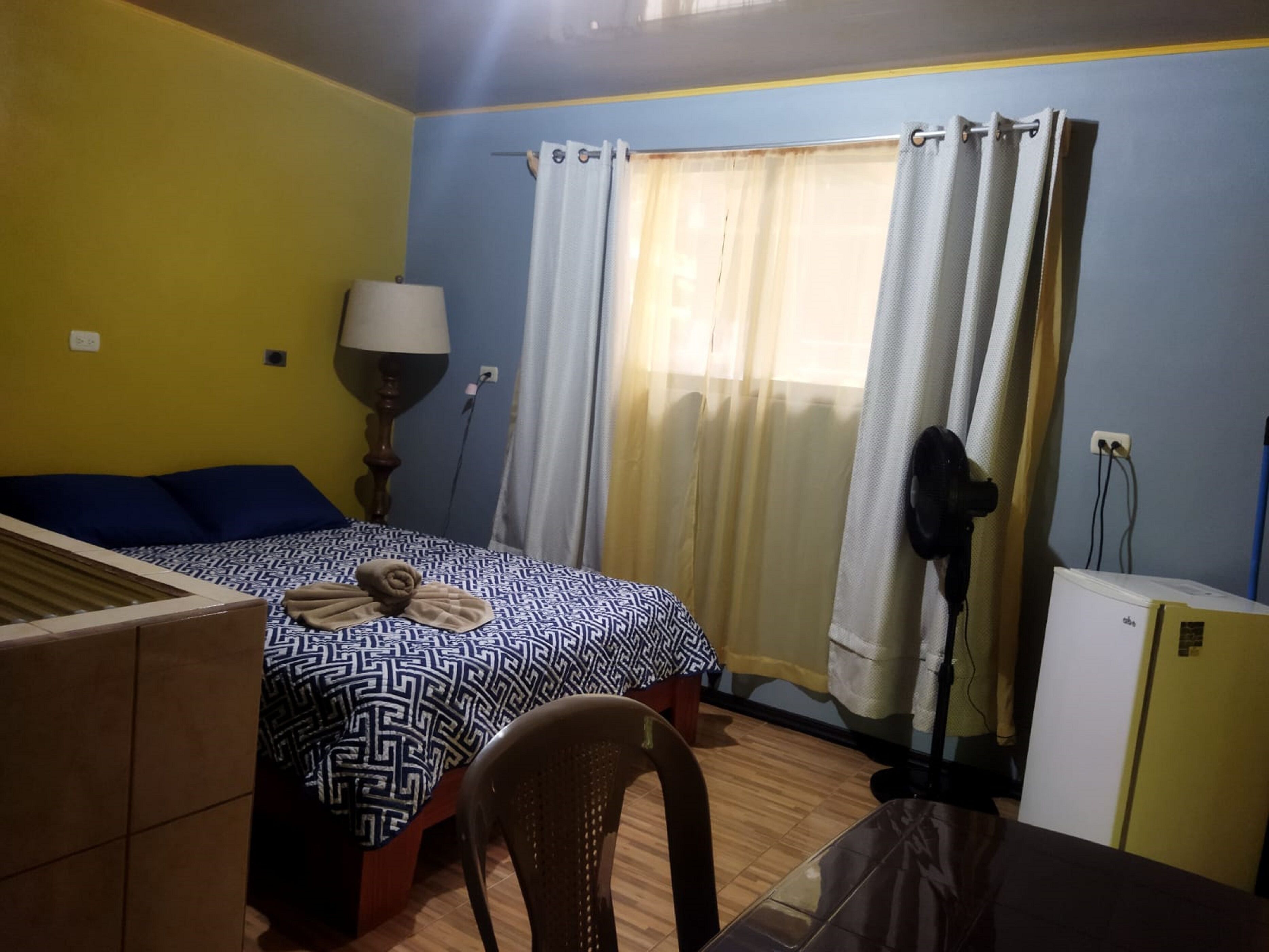Une pièce comprenant un lit, une chaise, un bureau, une lampe et une fenêtre avec des rideaux.
