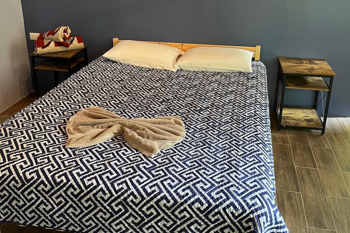 A neatly made bed with a geometric patterned bedspread, a single beige pillow, and a blanket draped over the side. A small wooden nightstand with a lamp and a red and white bag is beside the bed. A painting hangs on the wall above the bed.