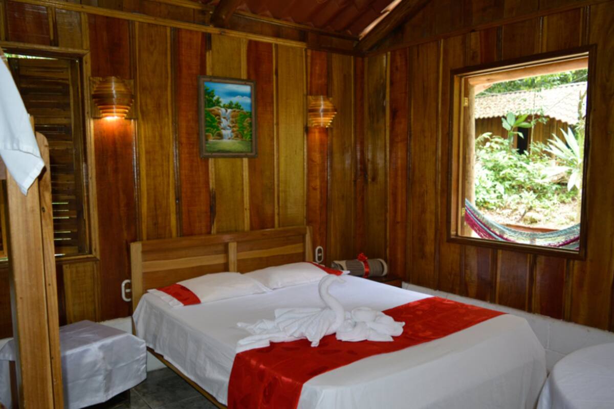 A wooden room with two beds, a painting on the wall, and a window with a view of greenery.