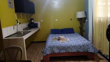 A compact room with a bed, a small desk with a sink, a chair, and a lamp.