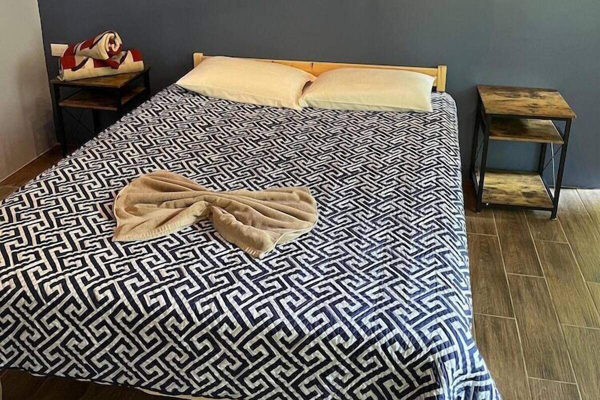A neatly made bed with a geometric patterned bedspread, a single pillow, and a blanket folded at the foot. A small wooden nightstand with a lamp and a red and white object on it is to the left. A painting hangs on the wall above the bed.