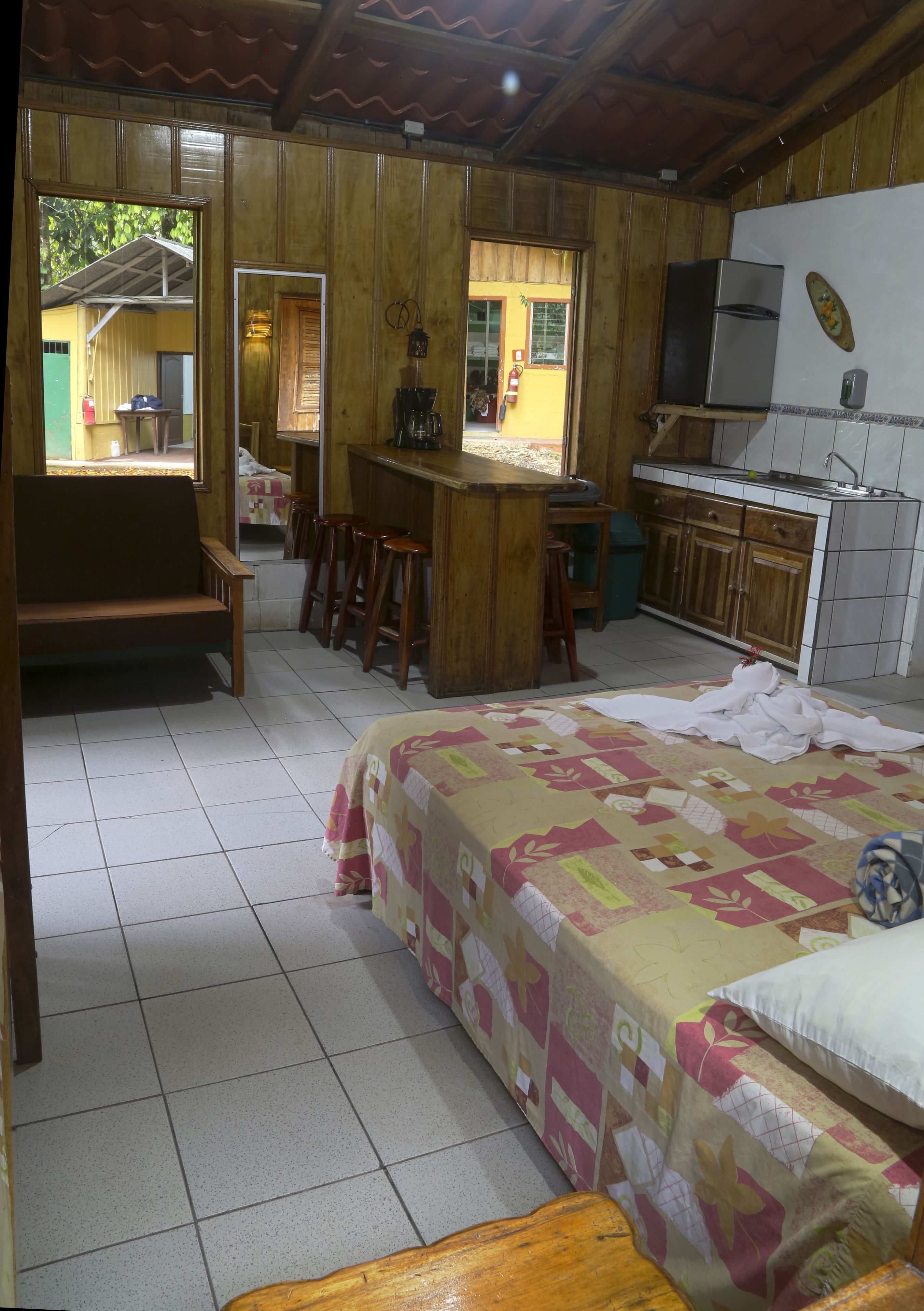 Una habitación acogedora con una cama, un banco de madera, una pequeña cocina con refrigerador y un comedor con taburetes.