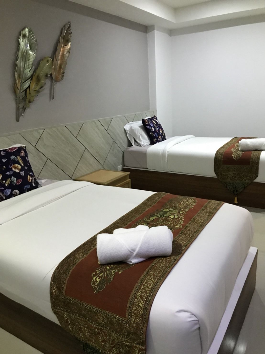 Ett hotellrum med två sängar, var och en med ett vitt överkast och en dekorativ röd och gul löpare. Mellan sängarna står ett nattduksbord med en lampa, och ovanför sängarna hänger ett dekorativt väggornament.