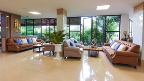 A spacious lobby with a brown leather sofa set, a large potted plant, and large windows offering a view of greenery. - Wish Hotel Ubon (Ubon Ratchathani)