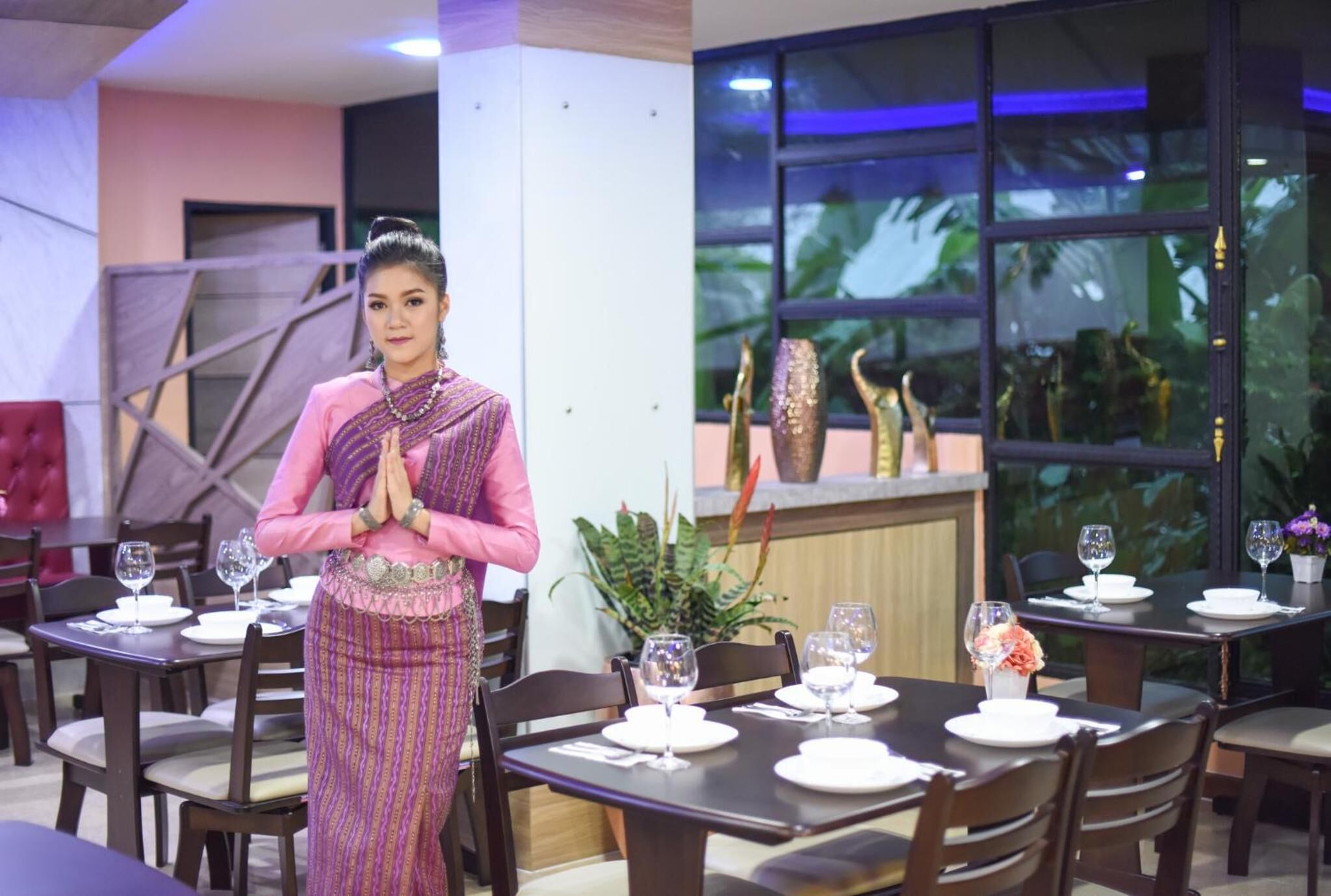 A woman in traditional attire standing in a dining area with tables set for dining, a large window with a view of greenery, and a modern interior design.