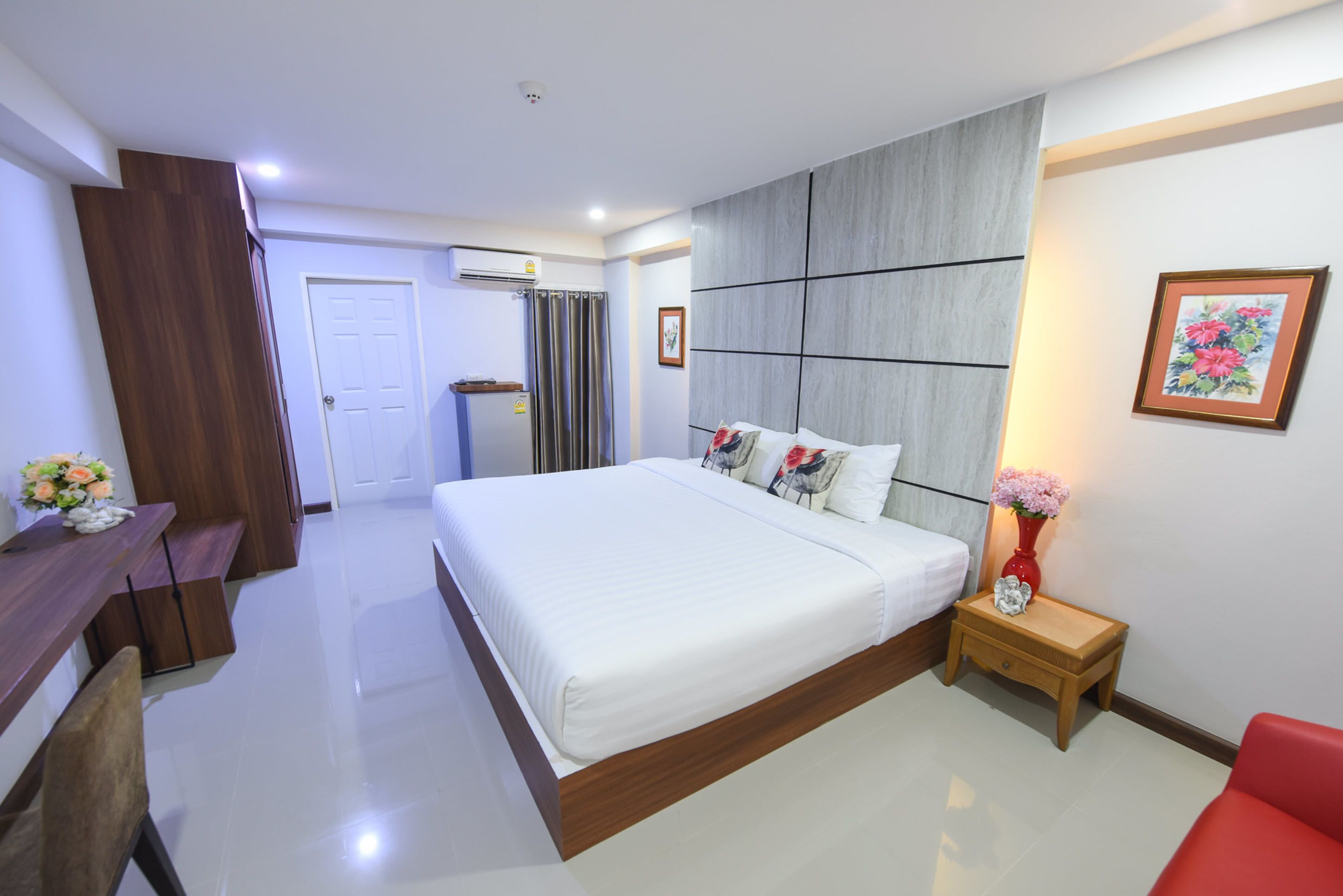A modern bedroom with a large bed, a wooden desk, a red chair, and a small bedside table with a vase.