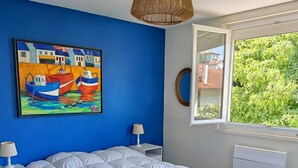 Une chambre avec un grand lit, un tableau représentant des bateaux, une fenêtre donnant sur un paysage verdoyant et un luminaire suspendu.