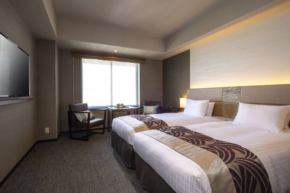 Une chambre d’hôtel avec un grand lit, un bureau avec une chaise, une télévision et une fenêtre avec des stores.