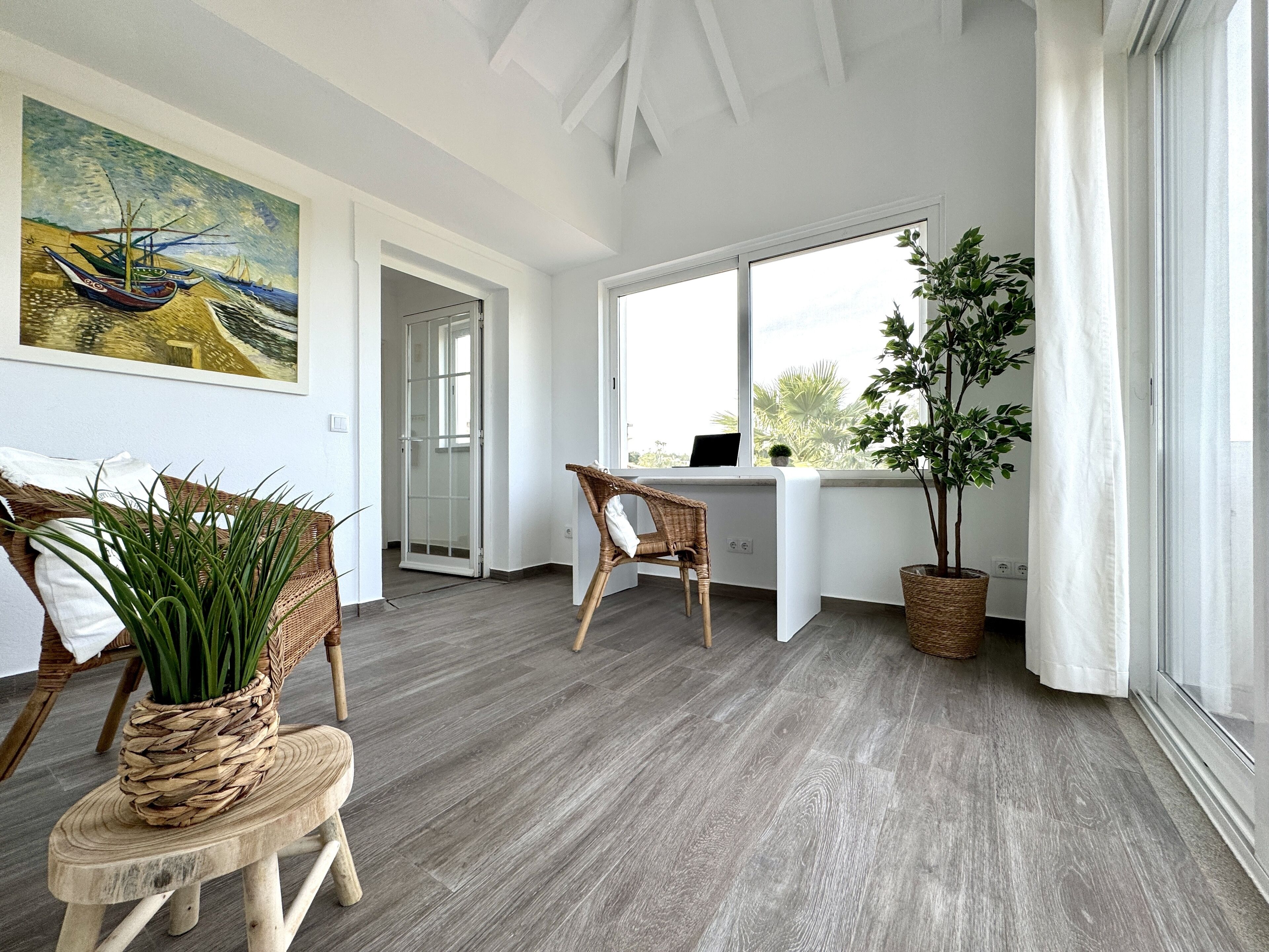 Une pièce lumineuse avec un plancher en bois, un bureau avec un ordinateur, une chaise, une plante et un tableau accroché au mur.