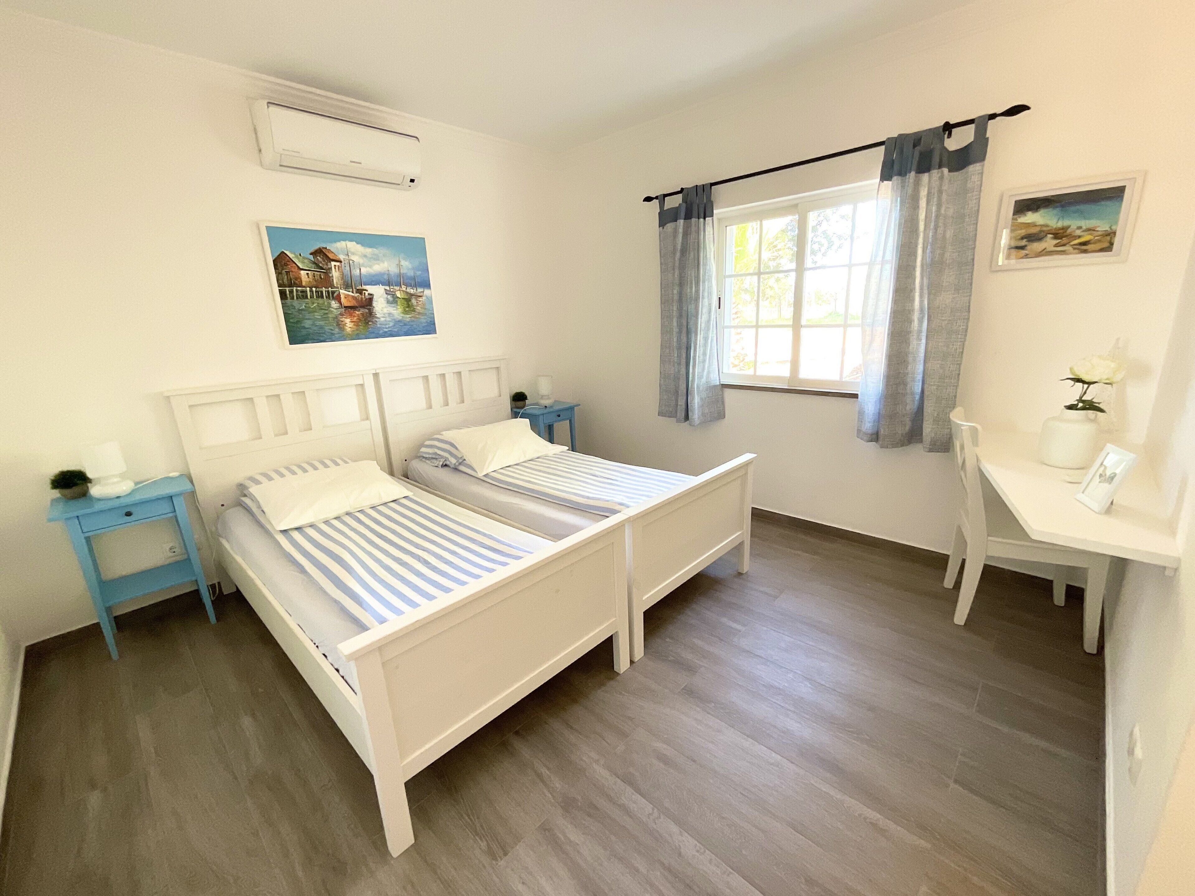 Une chambre avec deux lits simples, un bureau, une chaise, une table de chevet, une fenêtre avec des rideaux et un tableau au mur.