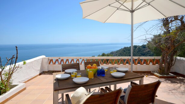 Un espace de restauration en plein air avec une table dressée pour deux personnes, un grand parasol et une vue sur la mer.