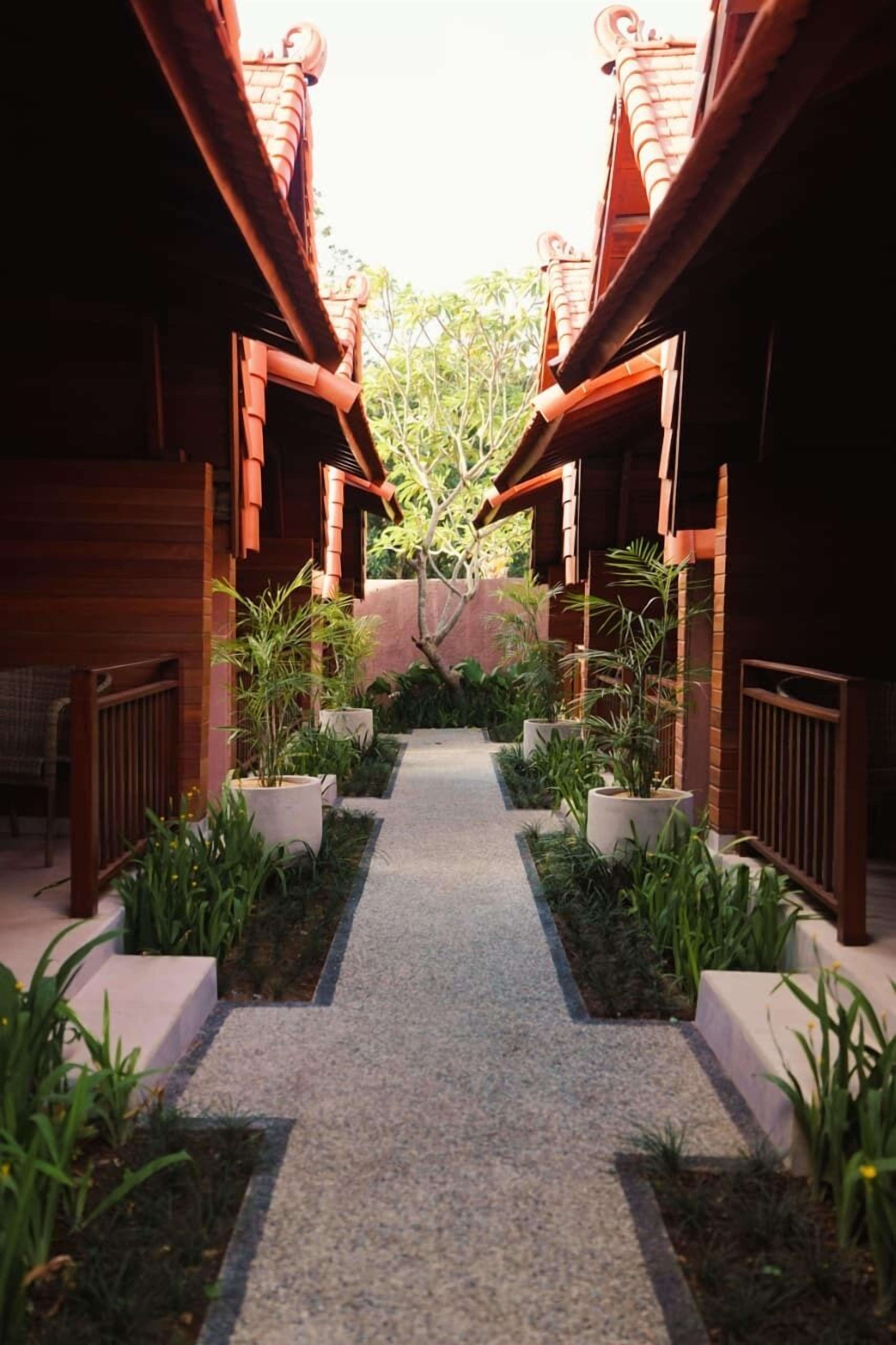 A pathway flanked by wooden structures with tiled roofs, leading to a central area with potted plants and a view of greenery.