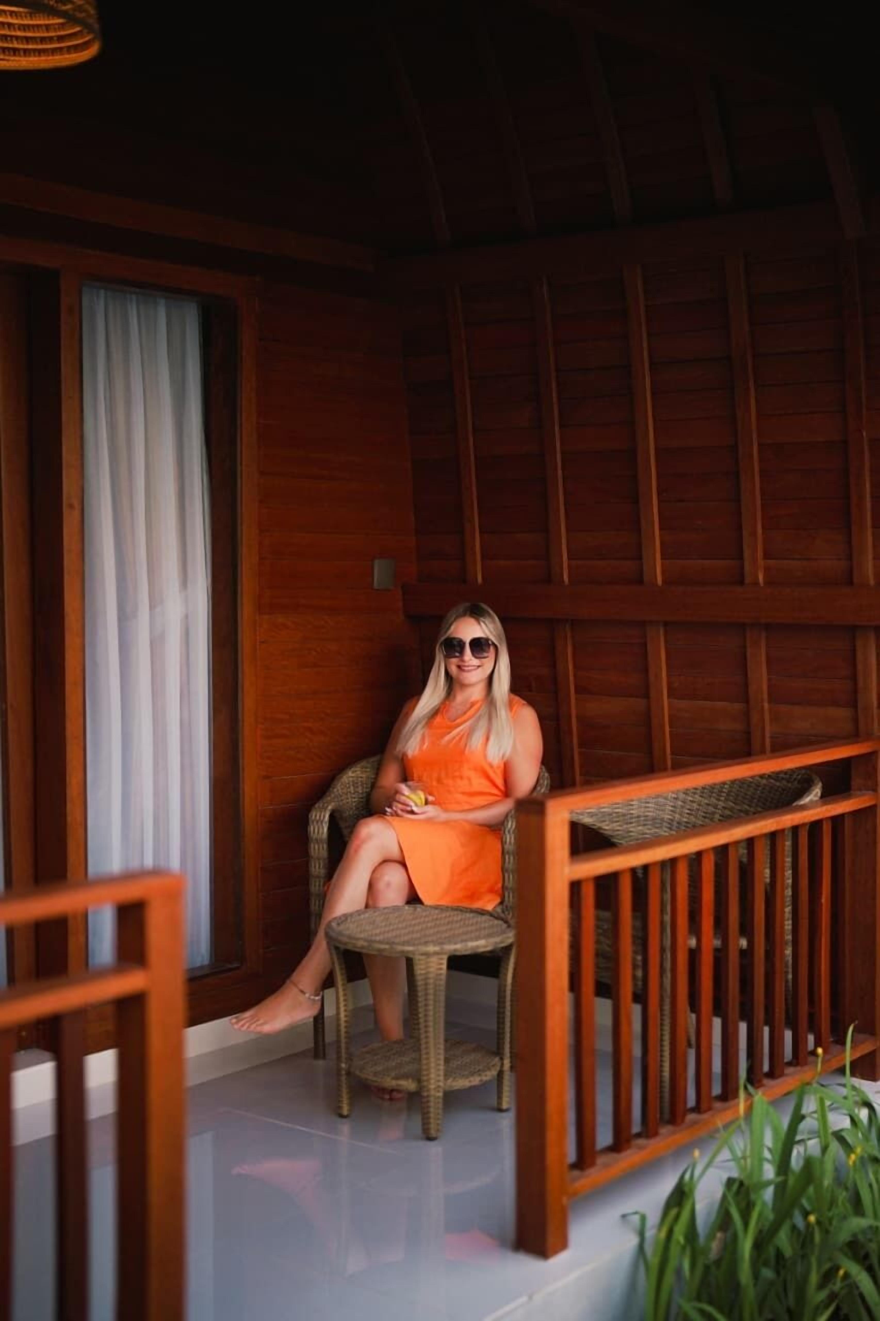 A person seated on a wicker chair in a wooden room with a glass floor, a wooden railing, and a window with white curtains.