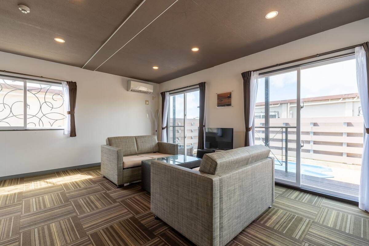 Un salon moderne avec un canapé, un fauteuil et une table basse en verre. On y trouve un téléviseur posé sur un meuble, une fenêtre avec des rideaux et un balcon avec vue.