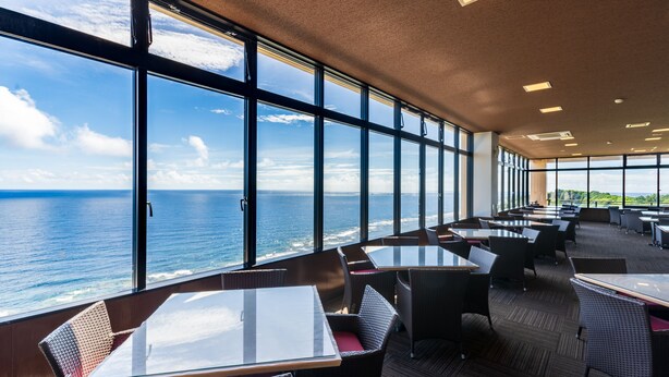 A restaurant with large windows offering ocean views, indoor seating with tables and chairs, and a ceiling with recessed lighting.