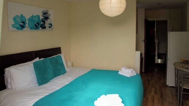 Une chambre d’hôtel avec un lit, un plaid turquoise, un oreiller blanc, un tableau au mur et un luminaire suspendu.