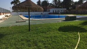 Un área de piscina con una estructura de techo de paja, césped y palmeras.