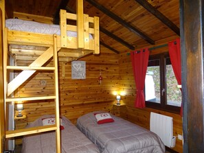 Quarto de cabana de madeira com duas camas, uma cama beliche, uma pequena mesa com abajur e uma janela com cortinas vermelhas.