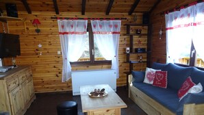 A cozy cabin interior with wooden walls, a stone floor, a wooden table with pine cones, a blue sofa with heart-patterned pillows, and a window with white curtains.
