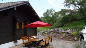A wooden cabin with a gravel patio, a red umbrella, and a stone wall with flowers.