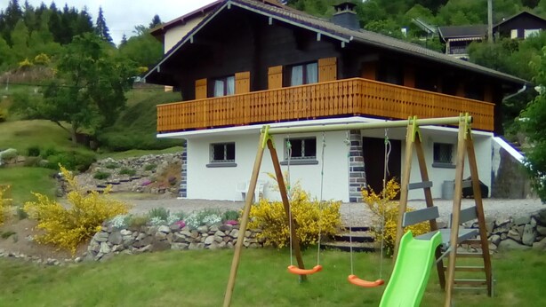 A house with a wooden balcony, a swing set, and a green slide in the yard.