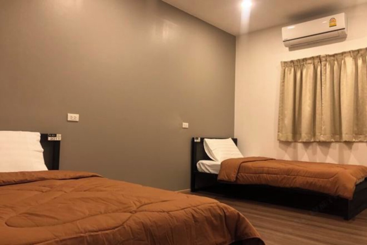 A hotel room with two beds, a window with curtains, an air conditioning unit, and a wall-mounted light switch.