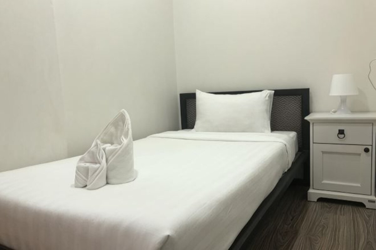 A neatly made bed with a white comforter and pillows, a small bedside table with a lamp, and a pair of shoes on the bed.