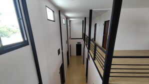 Un couloir doté d’un plancher en bois, de murs blancs et de rampes noires. Des fenêtres se trouvent sur le côté gauche et des portes sur le côté droit.