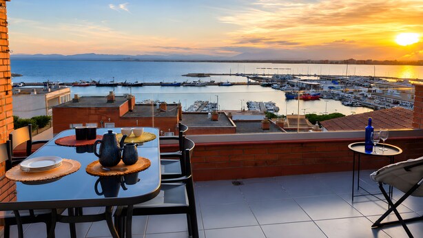 A terrace with a table set for dining, overlooking a marina with boats and a sunset view. - Golden Sunset Views - Panoramic Ocean View Duplex (Roses)