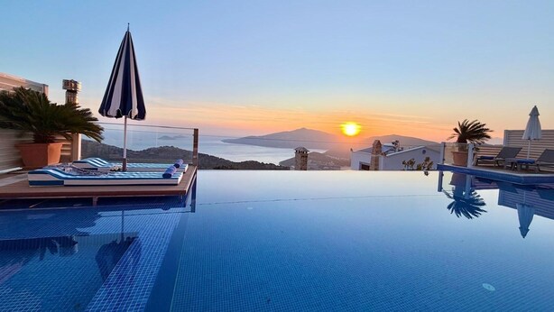 Una piscina infinita con sillas de descanso y sombrillas, vista al atardecer y un paisaje montañoso.