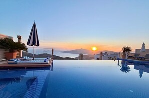 Una piscina infinita con sillas de descanso y sombrillas, vista al atardecer y un paisaje montañoso.