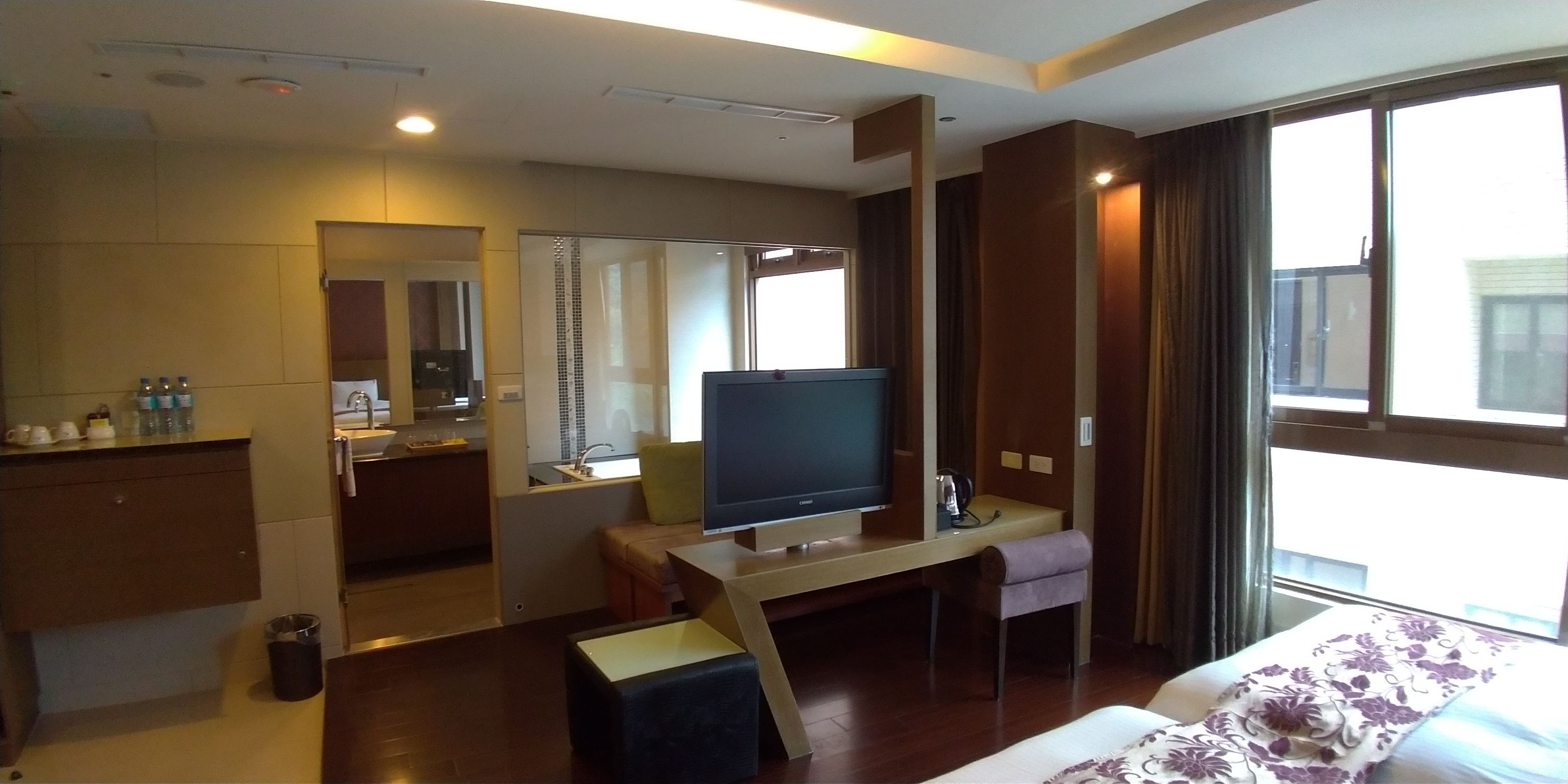 A hotel room with a bed, a desk with a television, a small table, a chair, a mirror, a window, and a kitchenette.