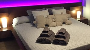 A neatly made bed with pillows and rolled towels, two bedside tables with lamps, and a purple ambient light strip above the bed.