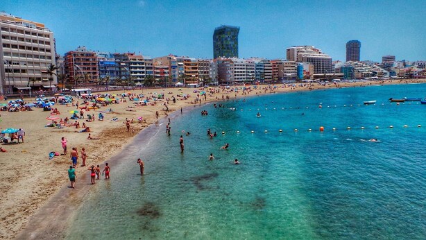 A beach with clear blue water, people swimming and sunbathing, and a cityscape in the background.