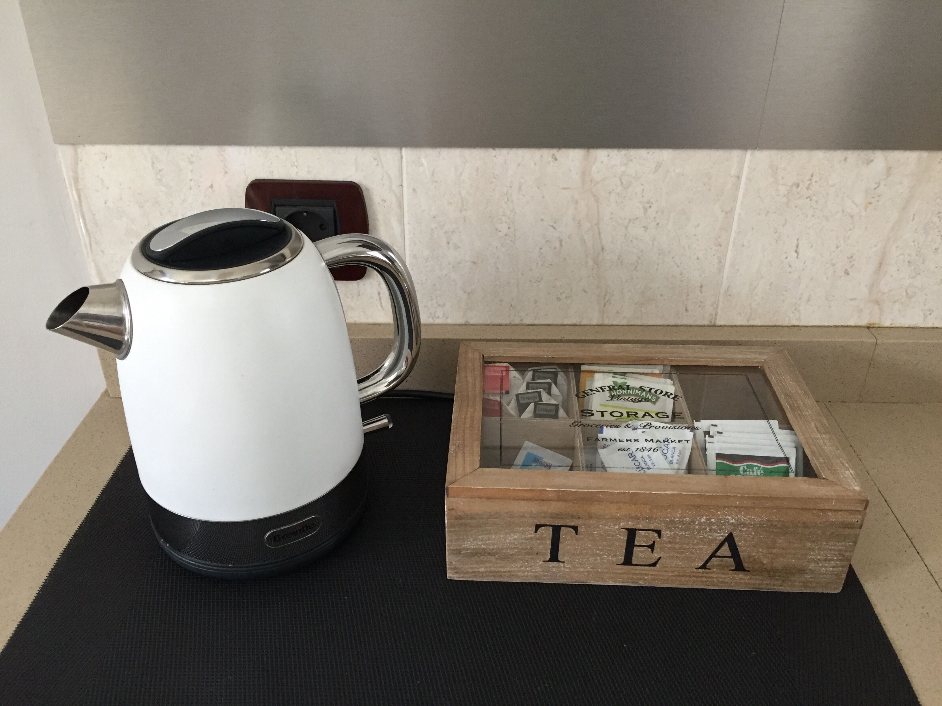 Una tetera eléctrica blanca y una caja de madera con la etiqueta 'TÉ' y bolsitas de té sobre una alfombra negra.