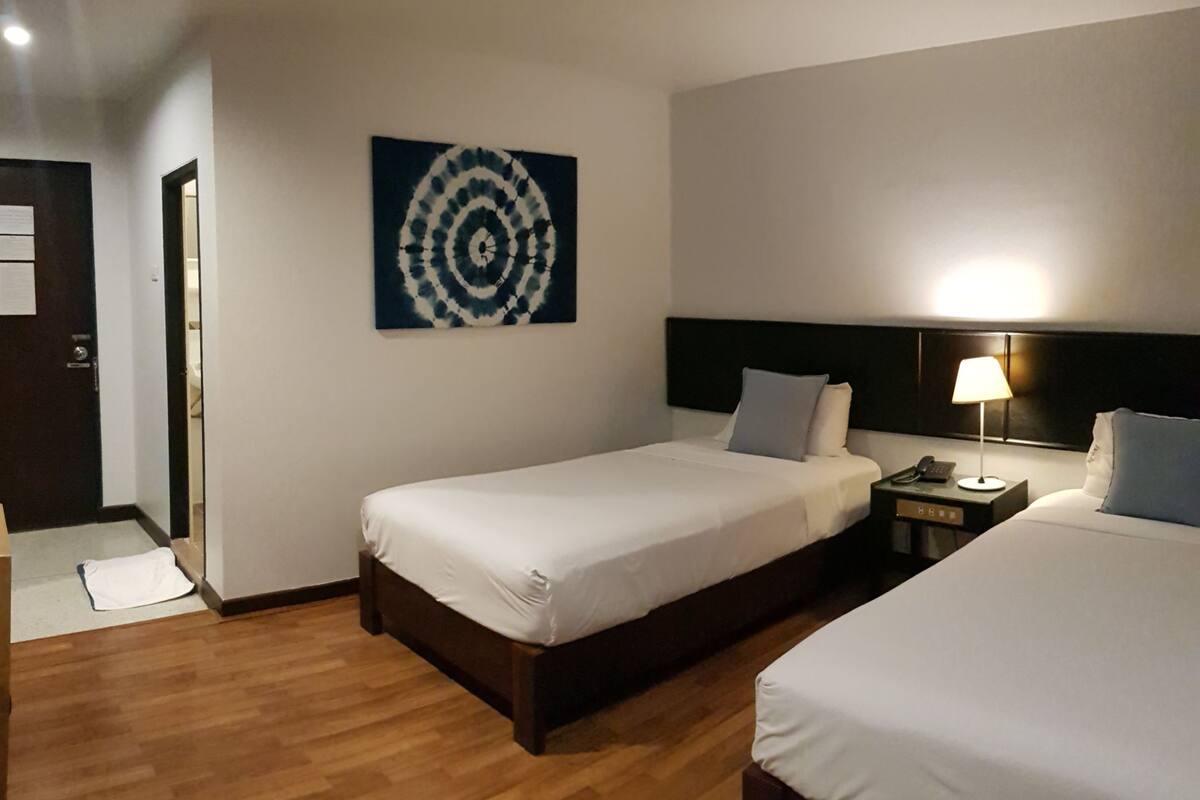 Habitación de hotel con dos camas individuales, piso de madera, un cuadro colgado en la pared, una mesita de noche con lámpara y una puerta con una toalla en el suelo.