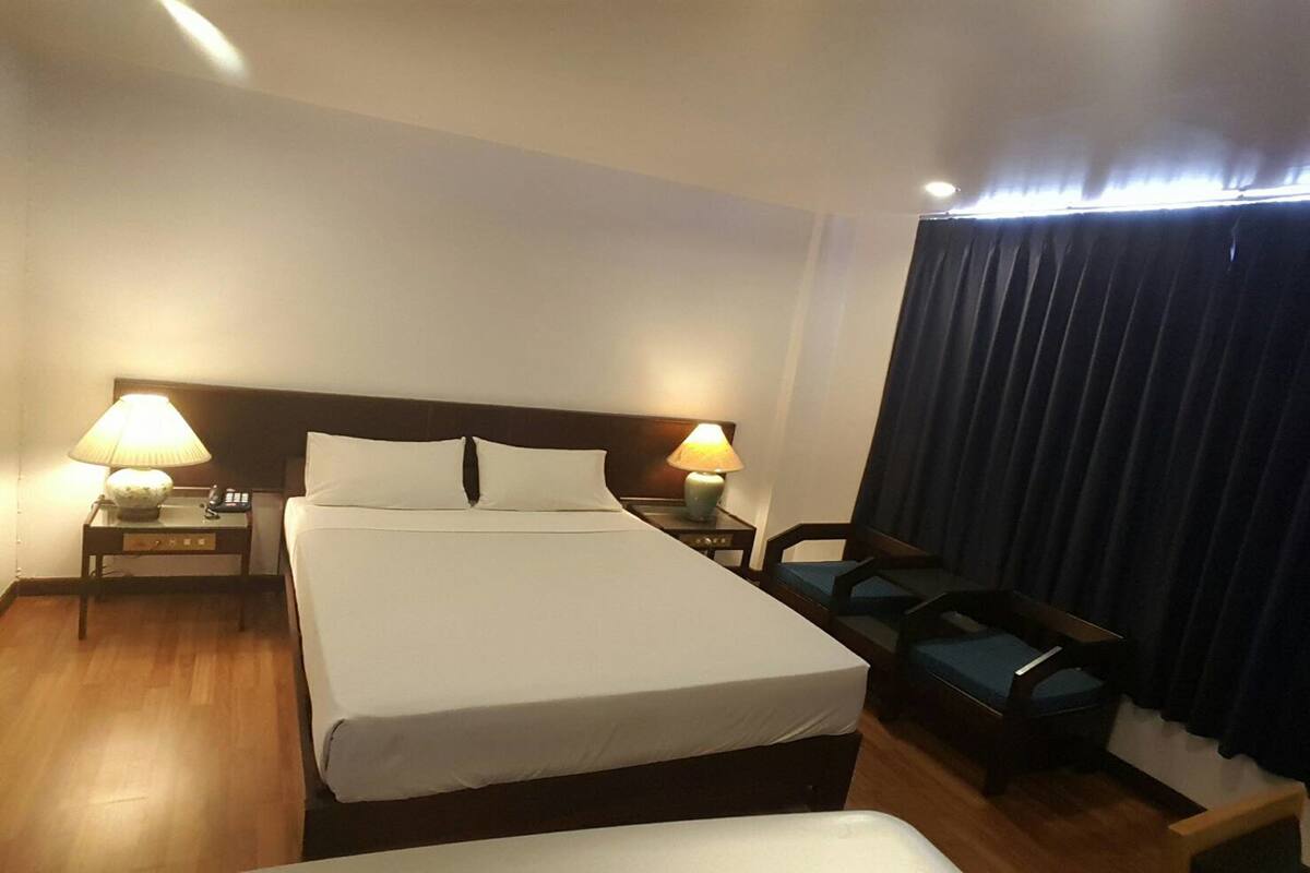 Una habitación de hotel con una cama grande, dos mesitas de noche con lámparas, una silla y una cortina.
