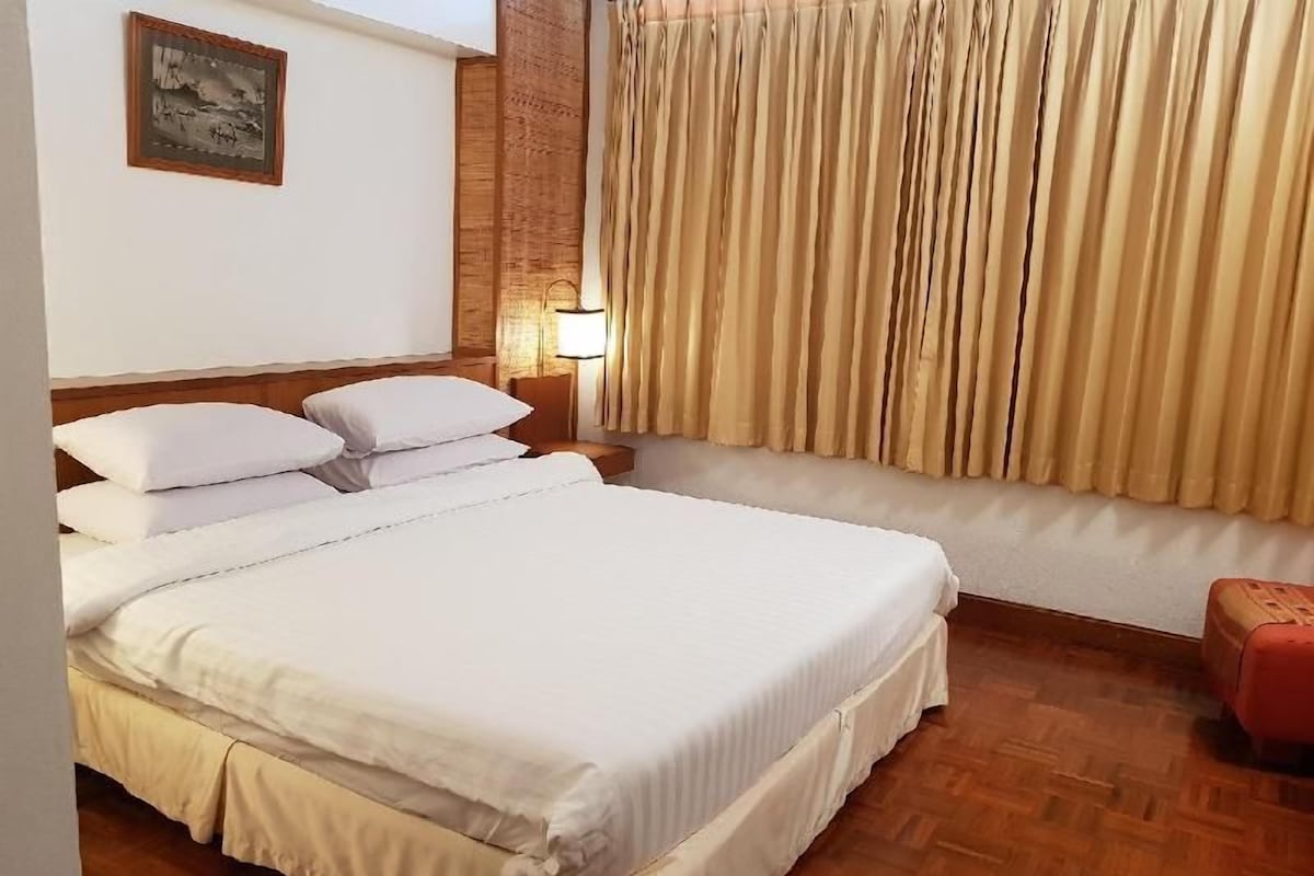 A hotel room with a bed, a nightstand, a lamp, a window with curtains, and a wooden floor.