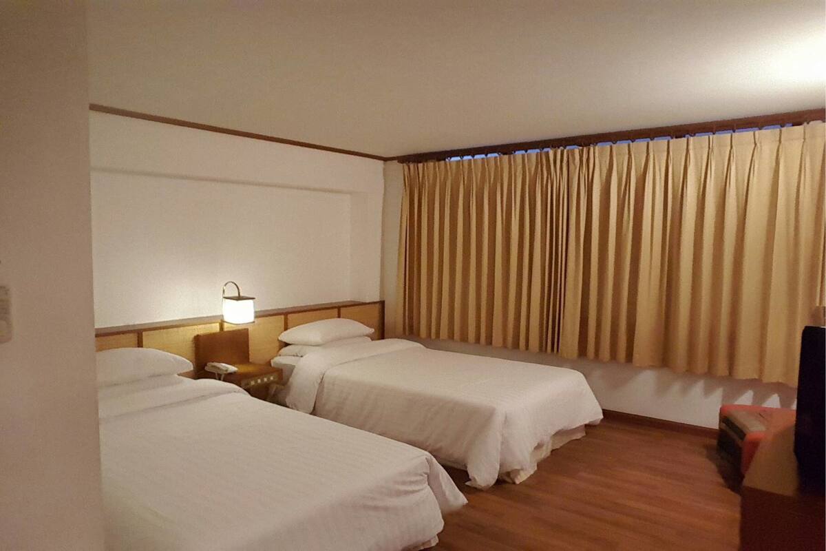 Habitación de hotel con dos camas individuales, una mesita de noche con lámpara, suelo de madera y una ventana con cortinas.
