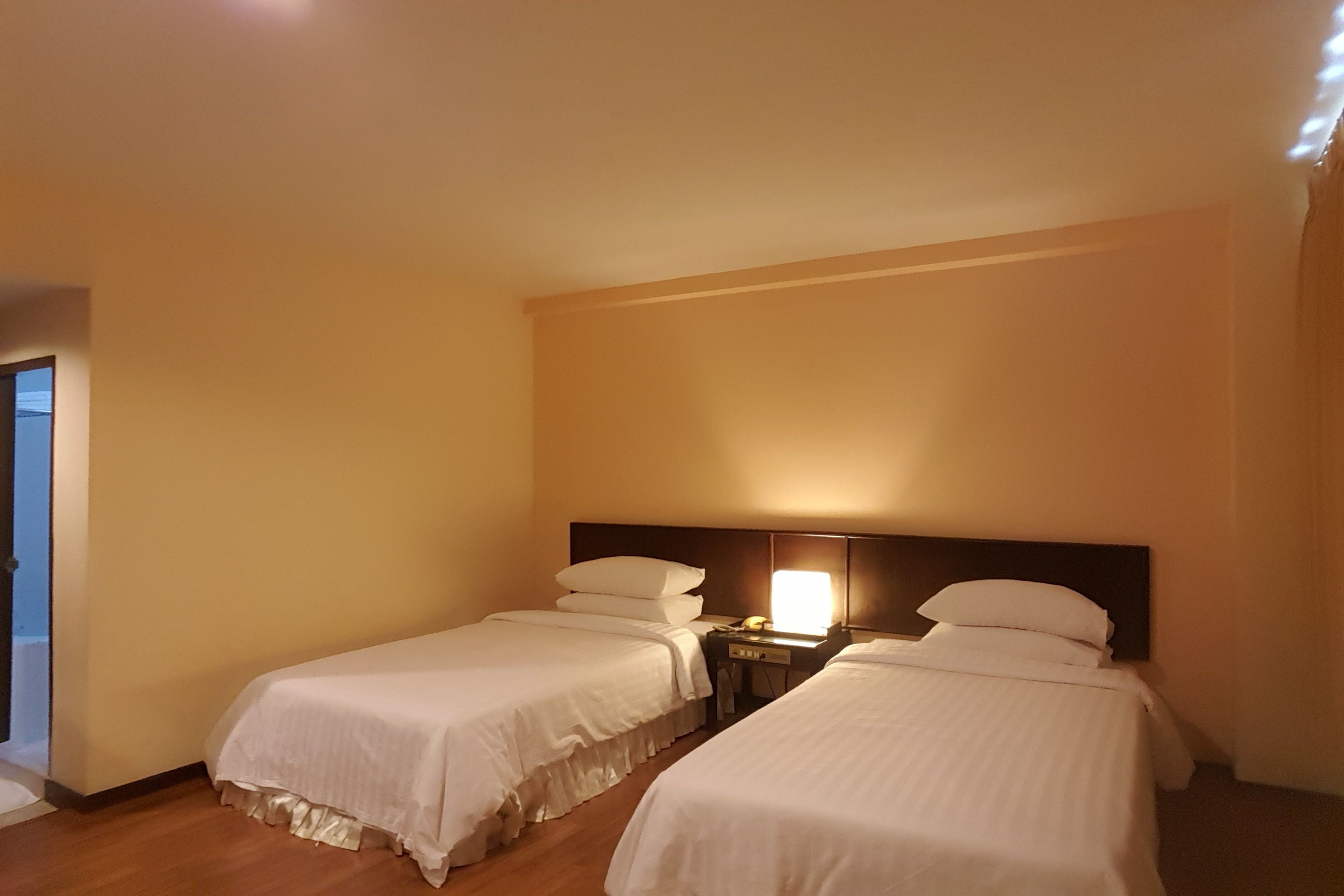Habitación de hotel con dos camas individuales, una mesita de noche con lámpara y vistas al exterior.