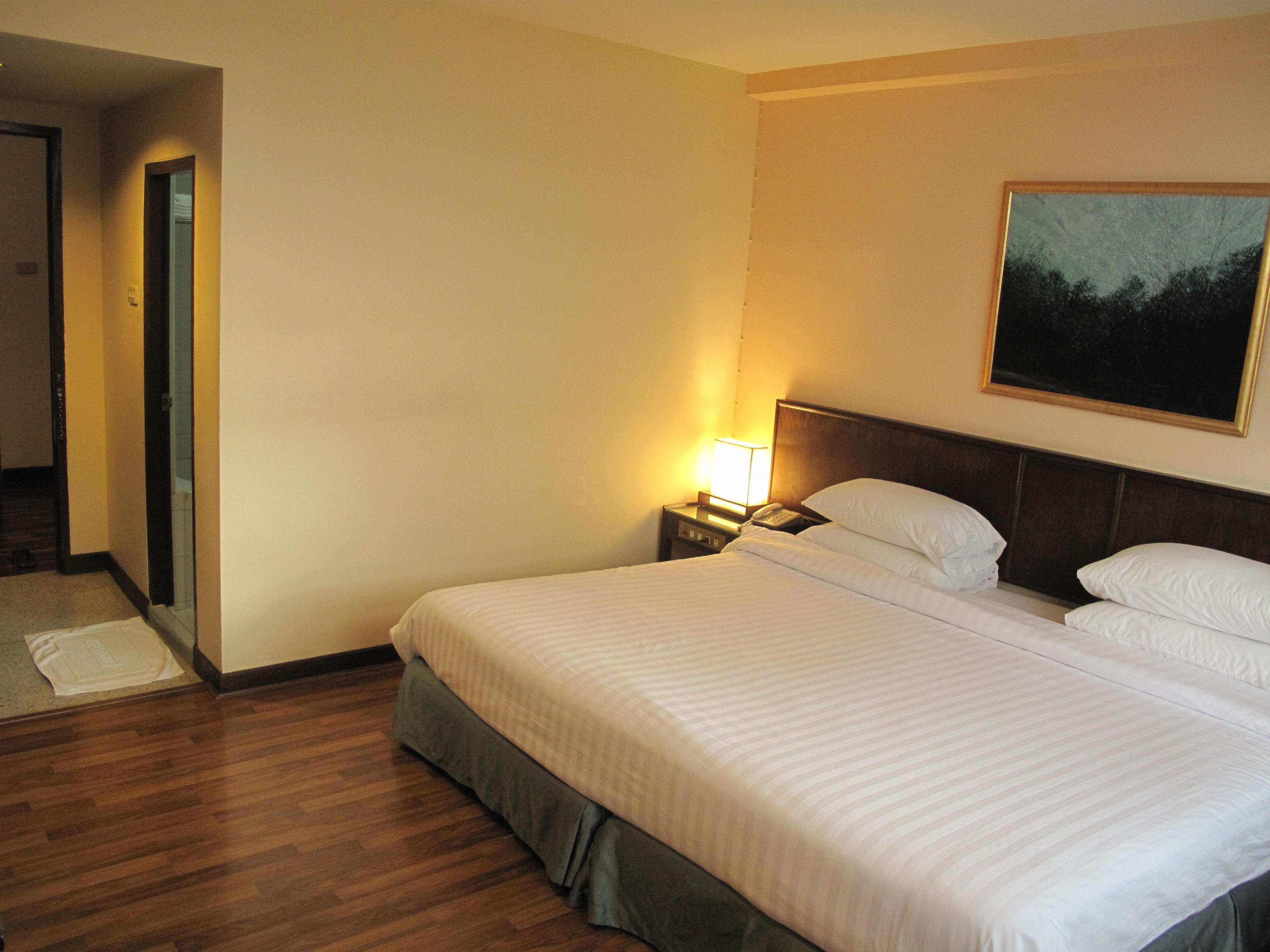 A hotel room with a large bed, a nightstand with a lamp, a painting on the wall, and a wooden floor.